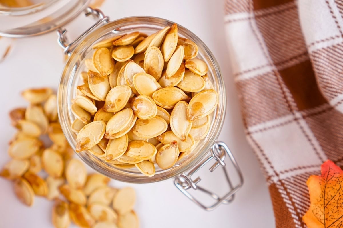 A jar filled with roasted pumpkin seeds next to a brown plaid cloth.
