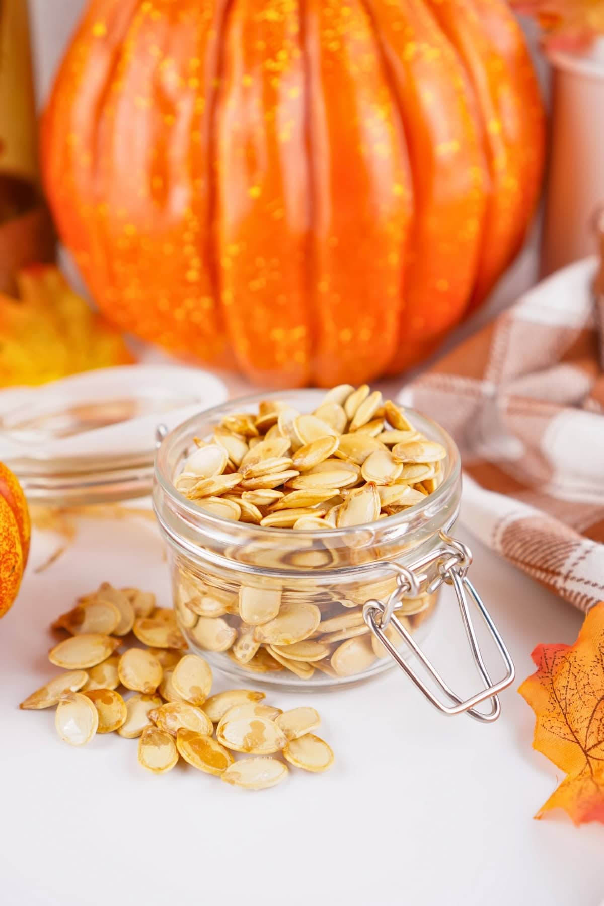 A jar of roasted pumpkin seeds in front of a large pumpkin and autumn decor.