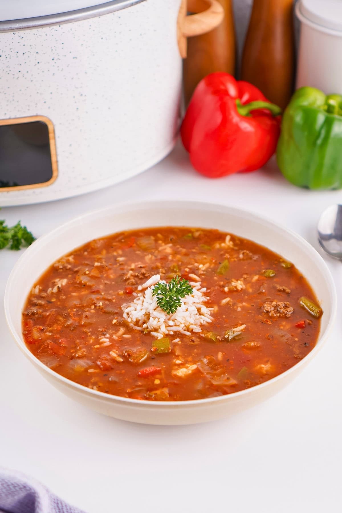 A bowl of soup with rice and parsley, with peppers and a slow cooker behind.