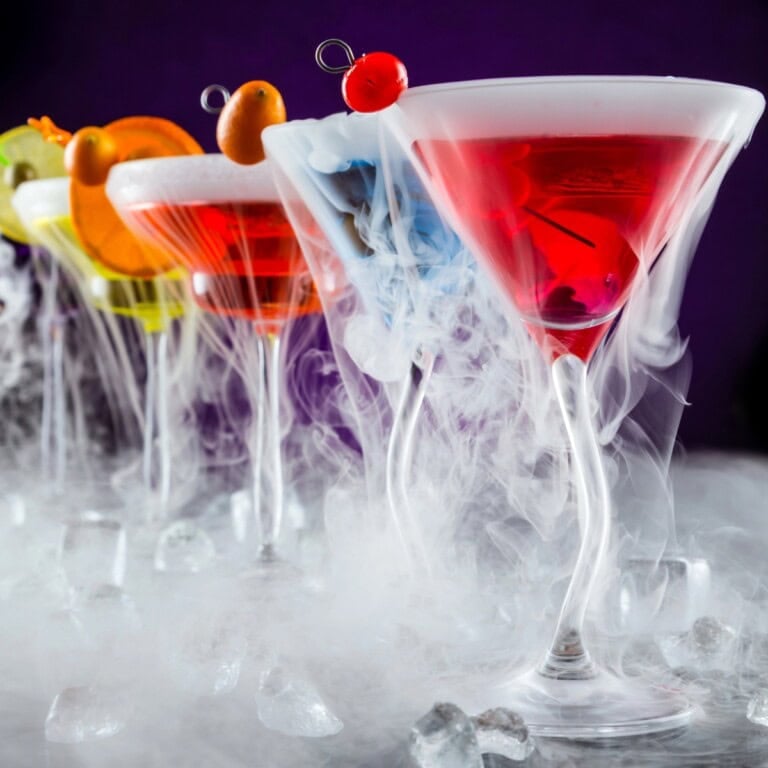 Four vibrant Dry Ice Cocktails in elegant glasses with smoke and creative garnishes.
