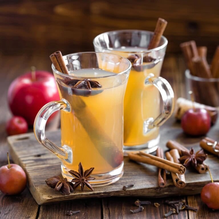 Two glasses of apple cider drinks with cinnamon sticks and star anise on a board.