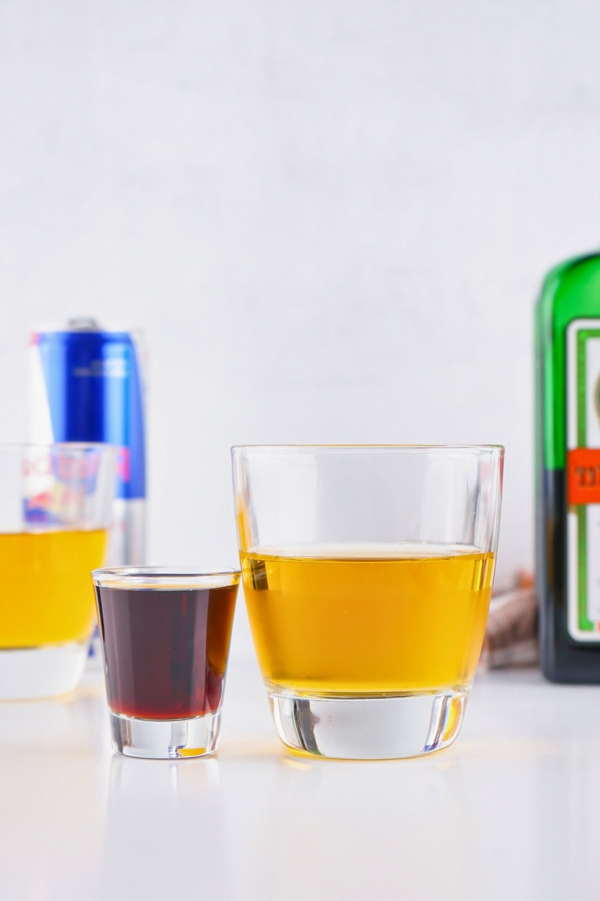 A shot glass of dark liquid and a glass of yellow drink on a white surface.
