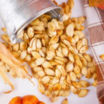 Oven baked pumpkin seeds spill from a metal bucket onto a white surface.
