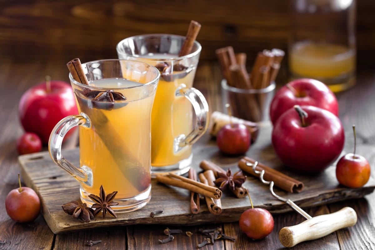 Two glass mugs of apple cider with cinnamon sticks and star anise, apples nearby.