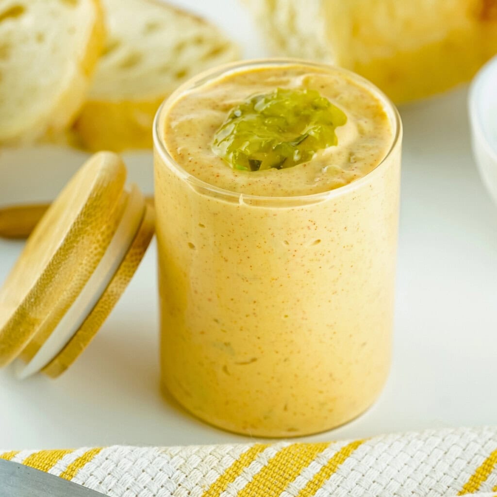 A glass jar of creamy sauce topped with relish, with bread slices in the background.
