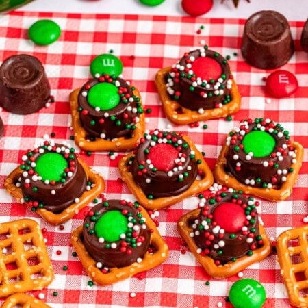 Chocolate pretzel treats with red and green candies and sprinkles on a checkered cloth.