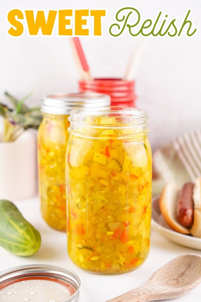 Jar of sweet relish on a table with a cucumber, hot dog, and kitchen items.