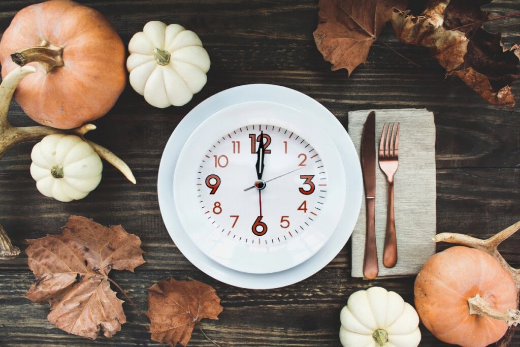 A clock on a plate surrounded by pumpkins, leaves, and cutlery on a table.