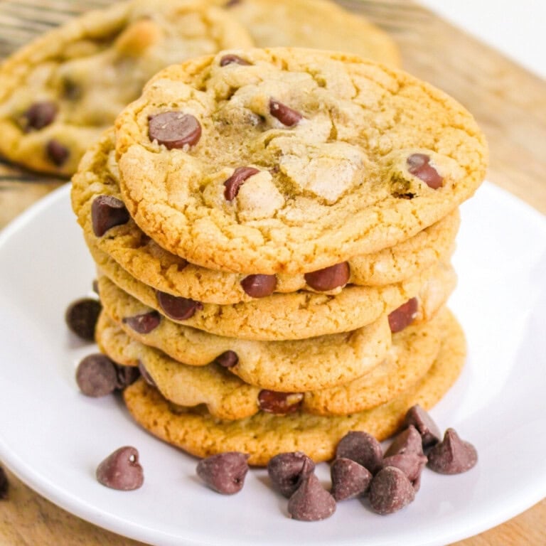 A stack of chocolate chip cookies on a white plate with extra chocolate chips.