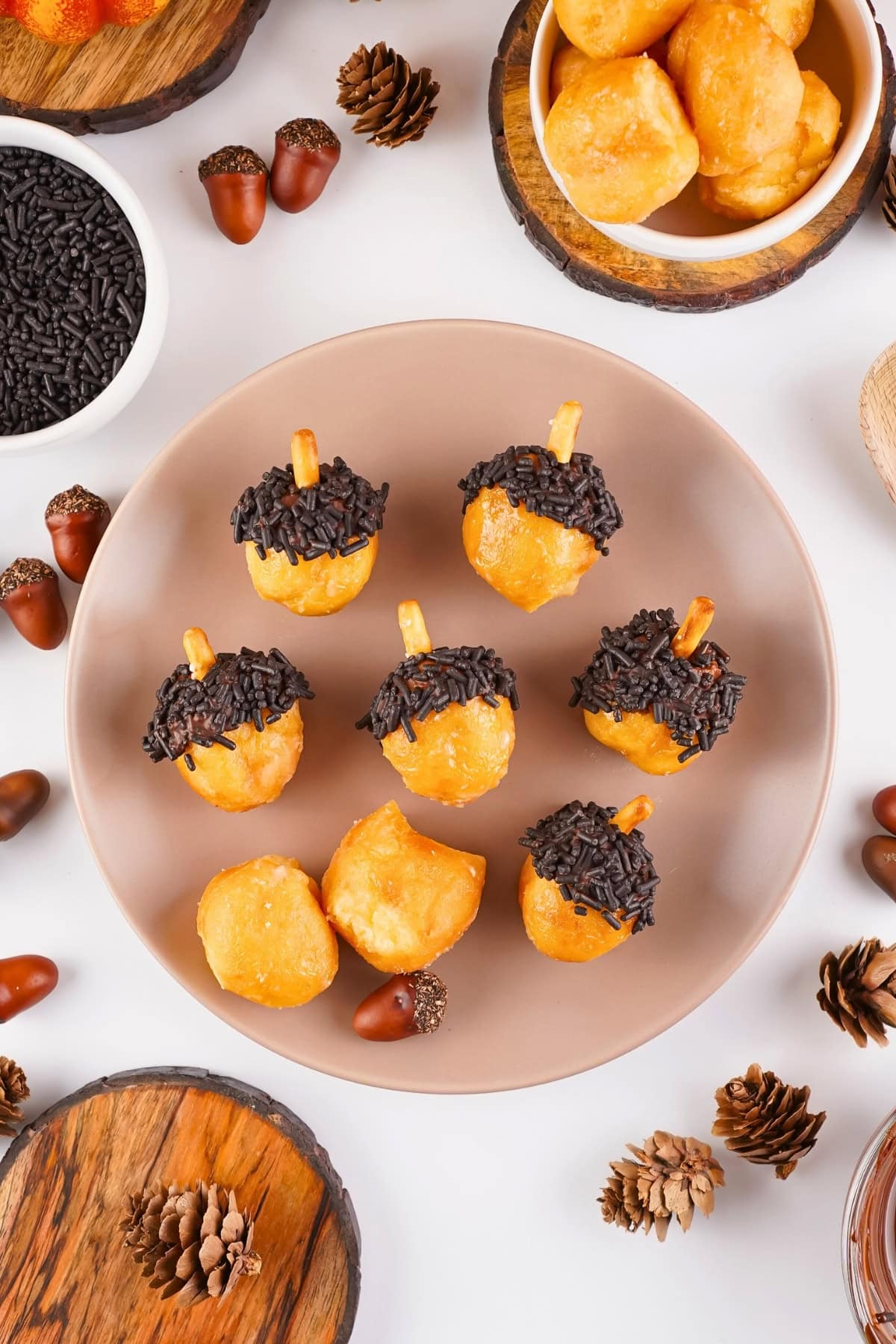 Donut holes decorated as acorns on a plate, surrounded by pinecones and acorns.