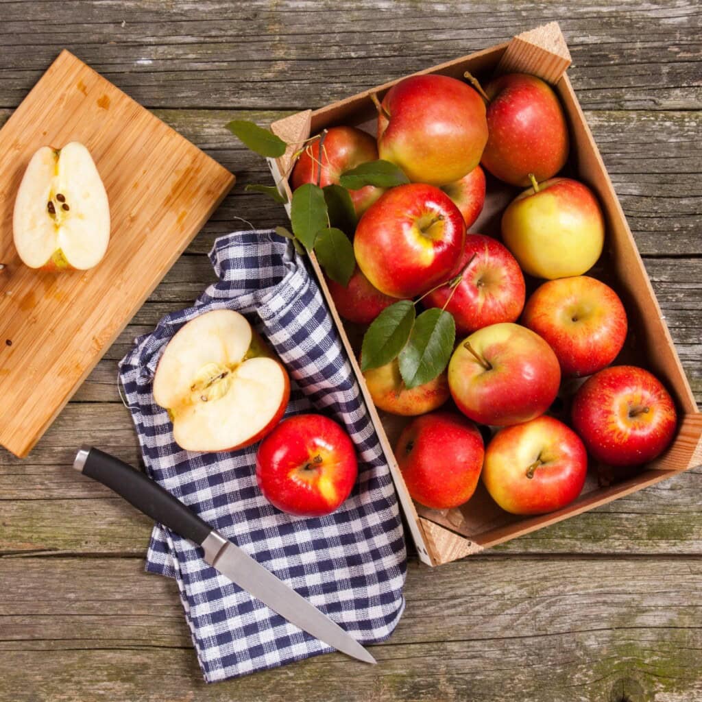 Red apples in a wooden box with cut apples, a knife, and a checkered cloth.