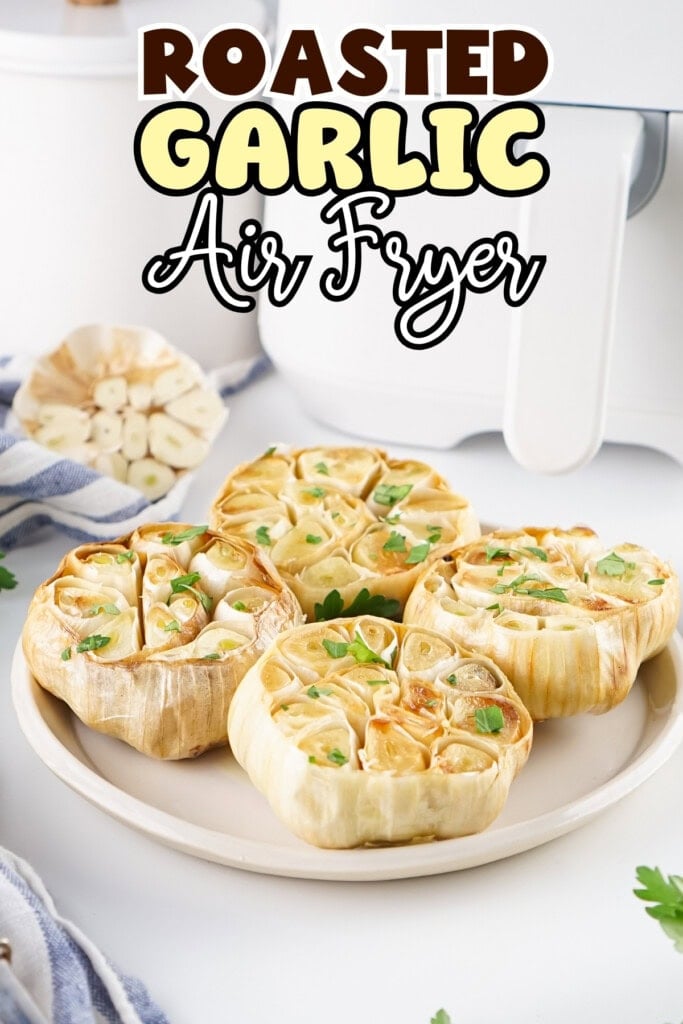 Four roasted garlic bulbs garnished with parsley on a white plate near an air fryer.