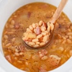 A wooden spoon lifts beans, ham, and broth from a white slow cooker.