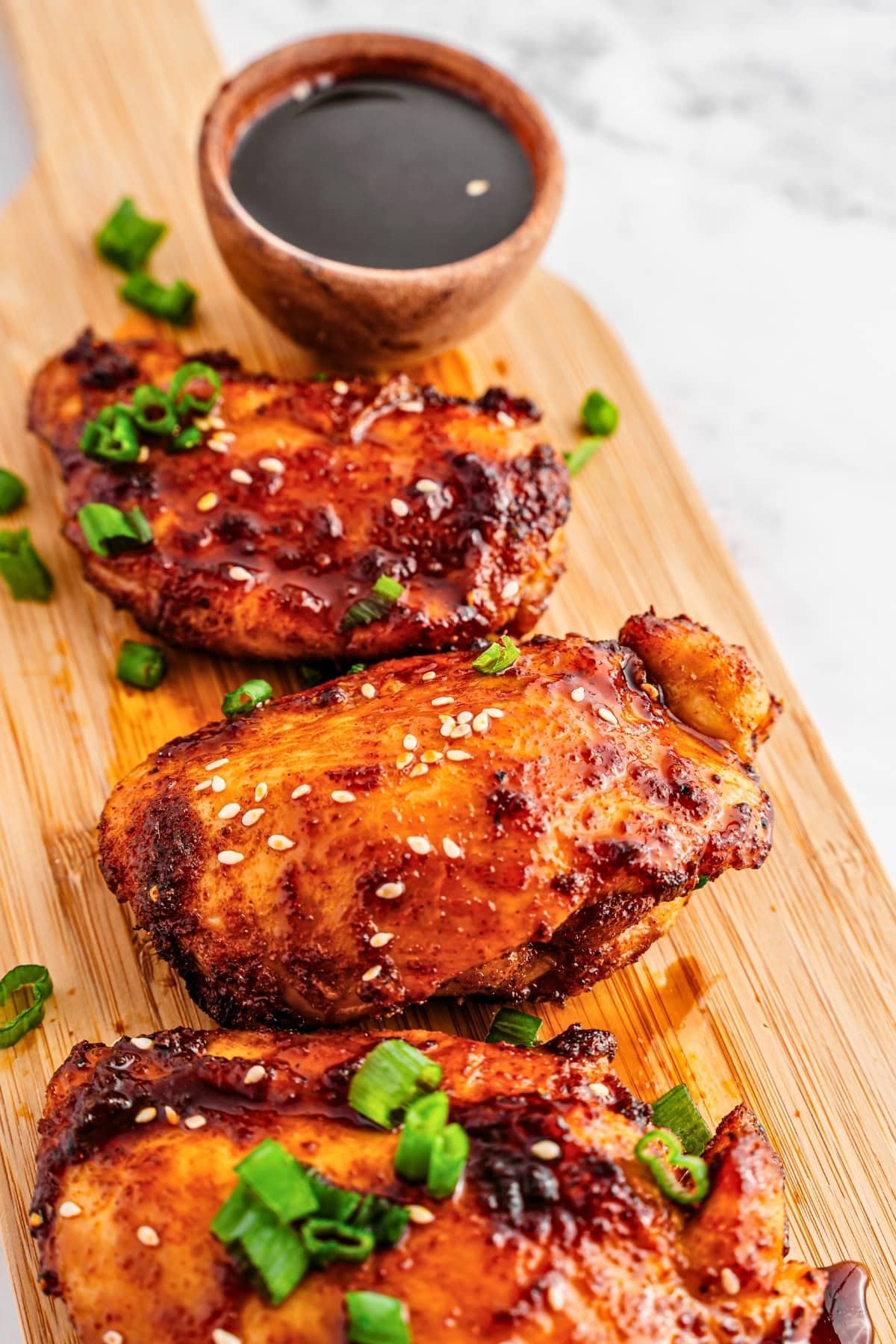 Grilled chicken thighs with sesame seeds, green onions, and dipping sauce on a board.