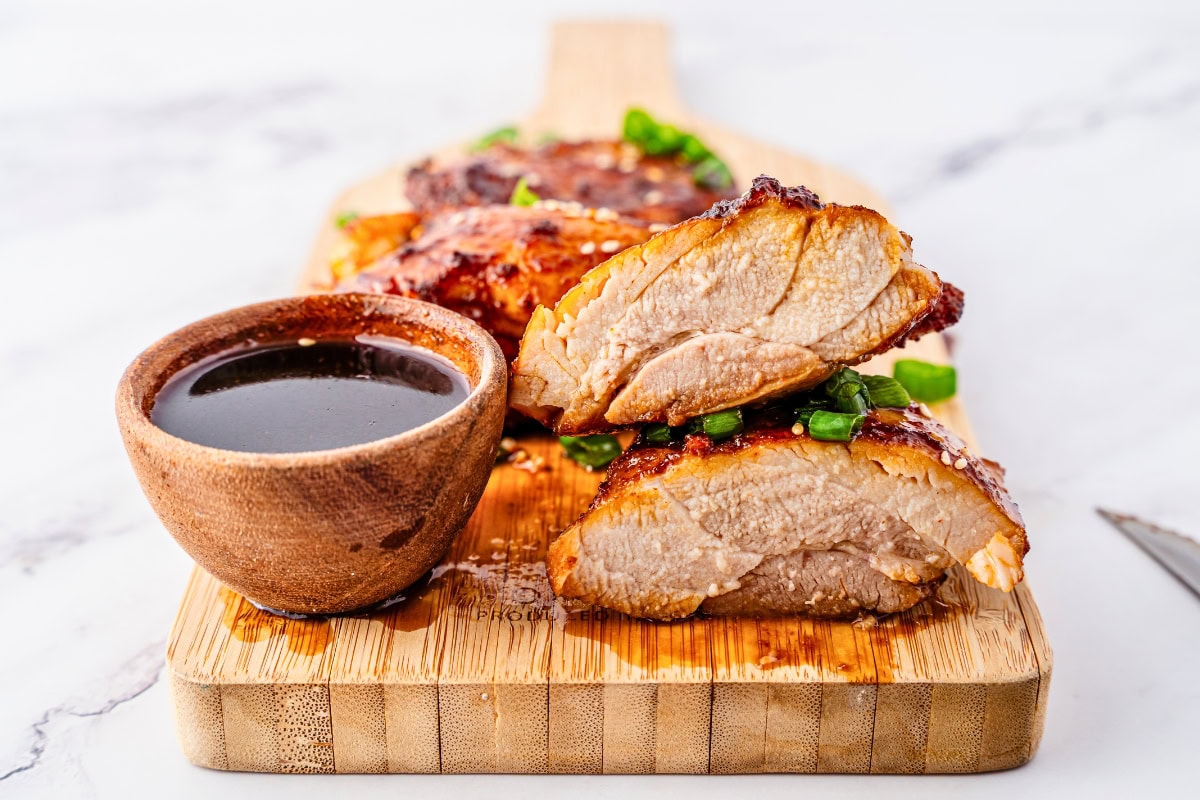 Sliced grilled chicken breast with sauce in a wooden bowl on a cutting board.