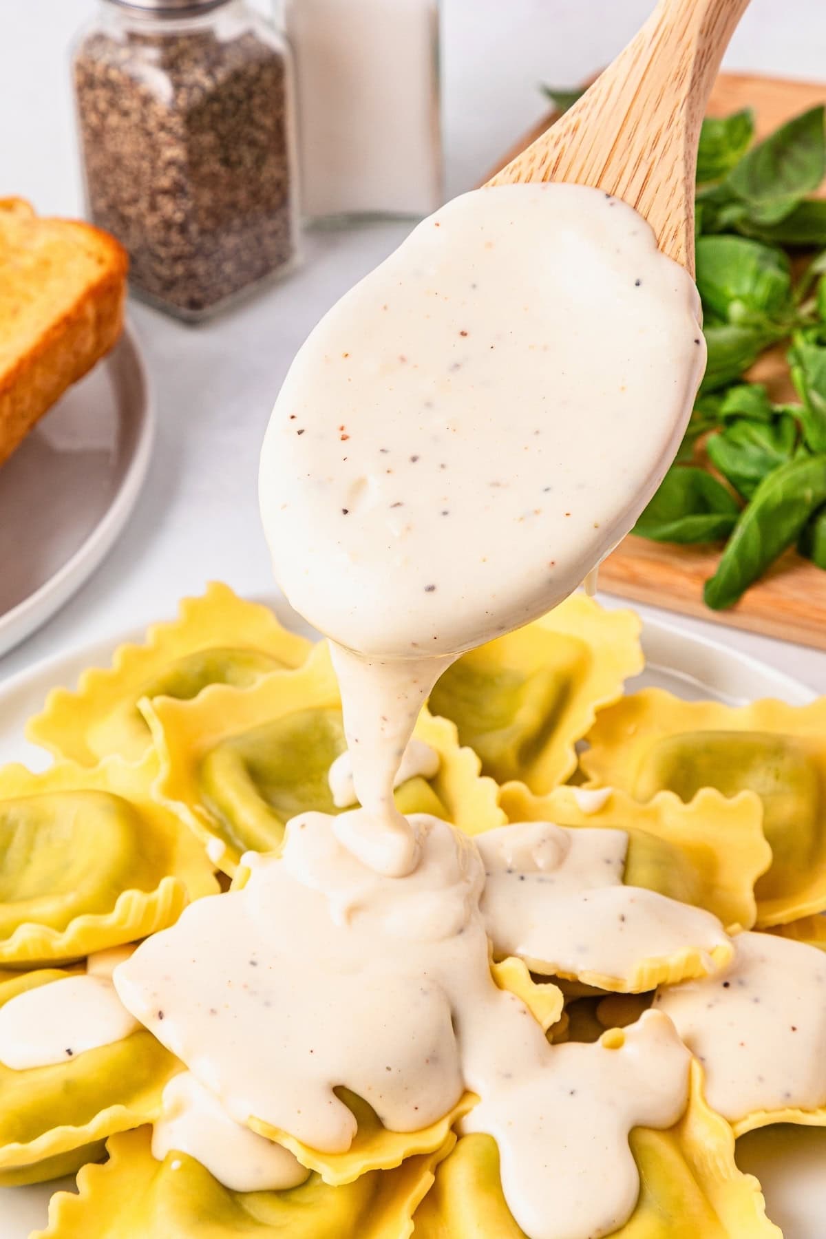 A creamy white sauce being poured over spinach ravioli on a plate.
