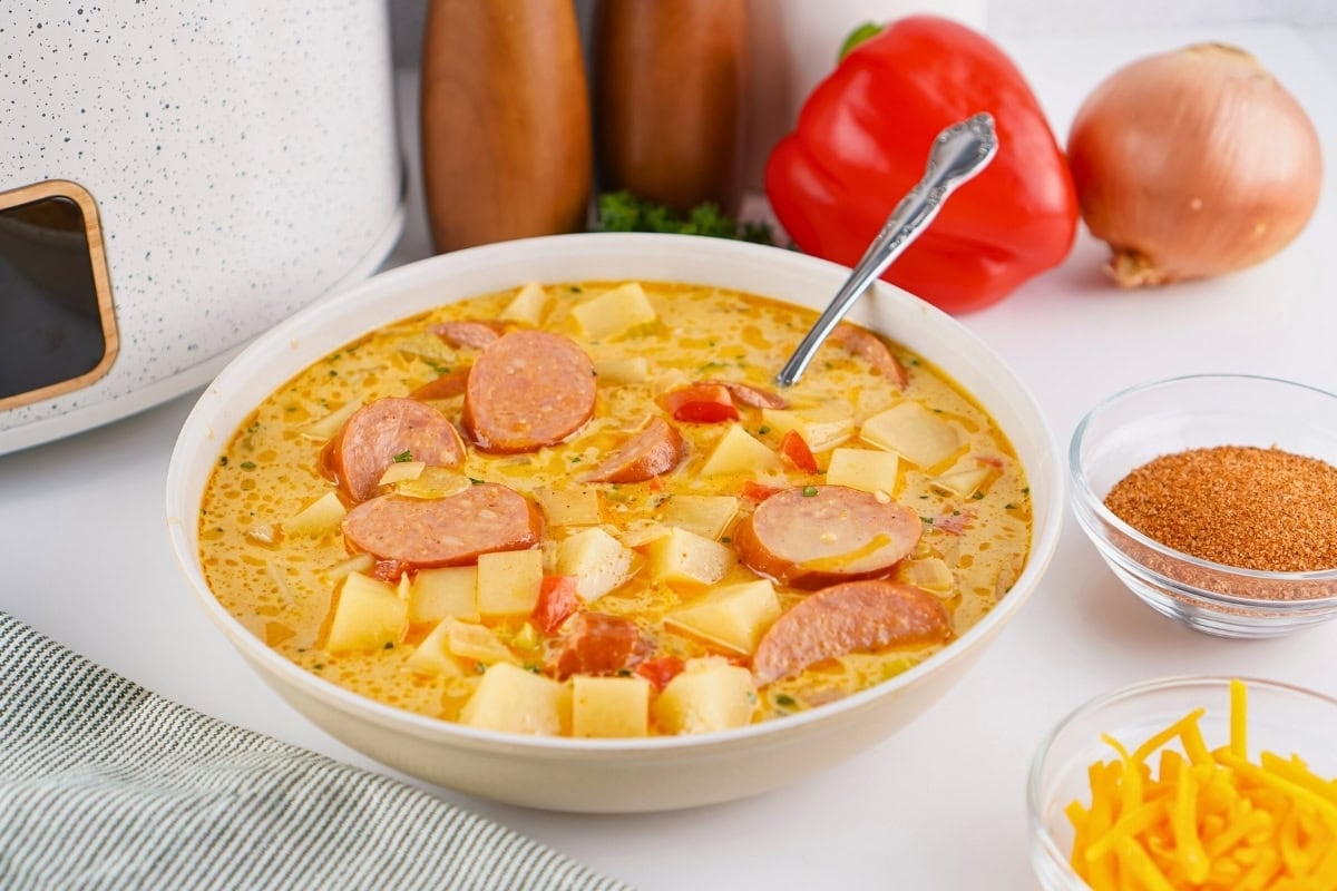 Bowl of creamy sausage and potato soup with a spoon, surrounded by ingredients.