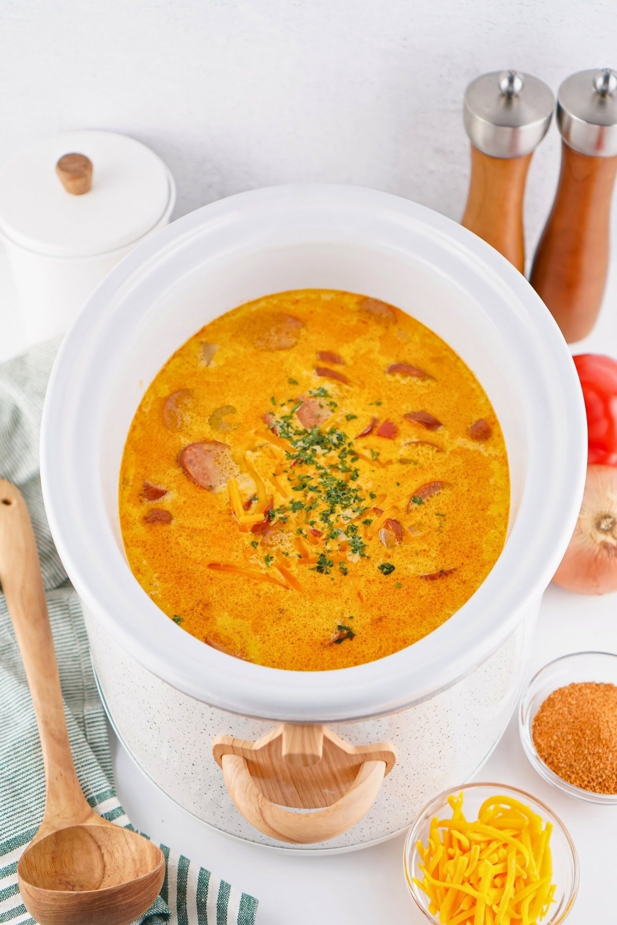 A slow cooker filled with cheesy sausage soup, surrounded by ingredients and utensils.