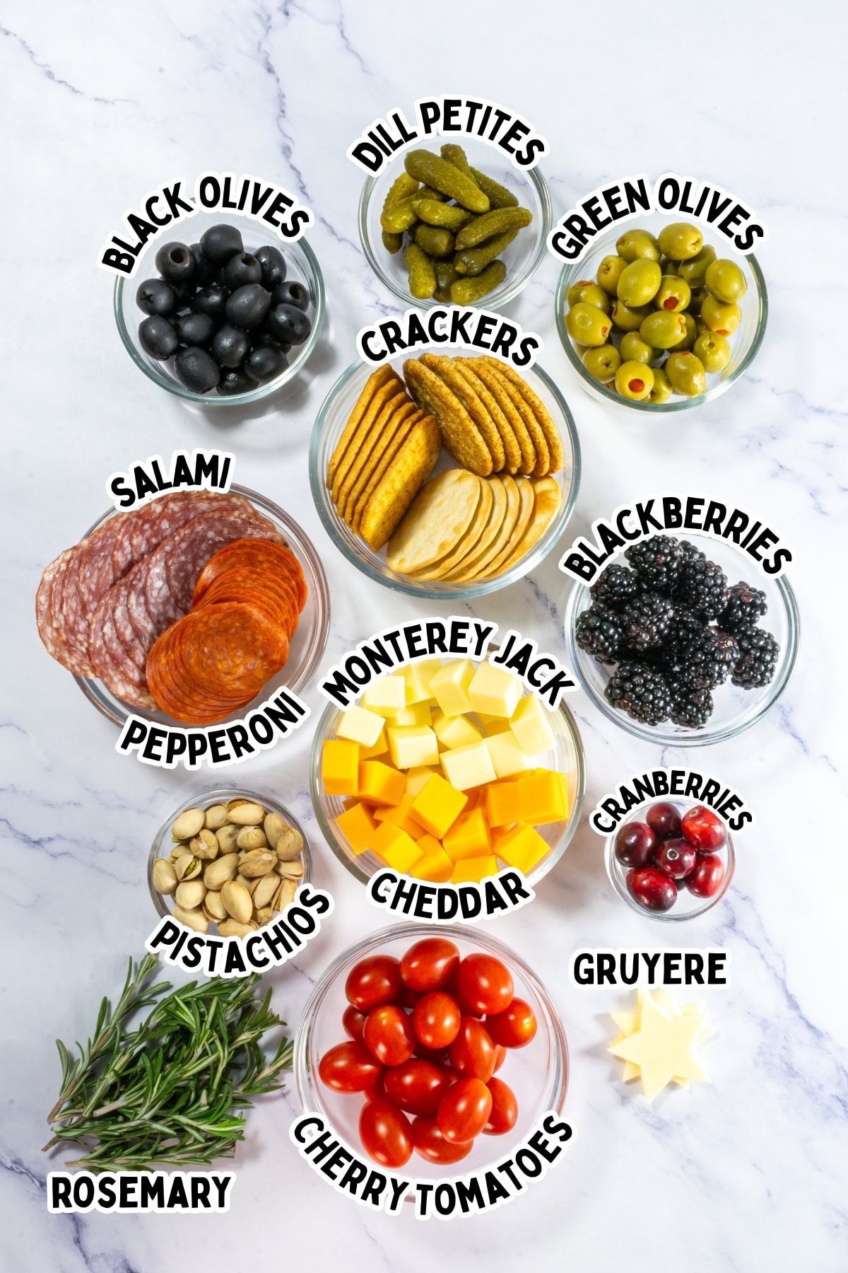 Assorted charcuterie ingredients labeled in glass bowls on a white marble surface.