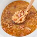 A wooden spoon scoops baked beans with bacon from a white bowl.