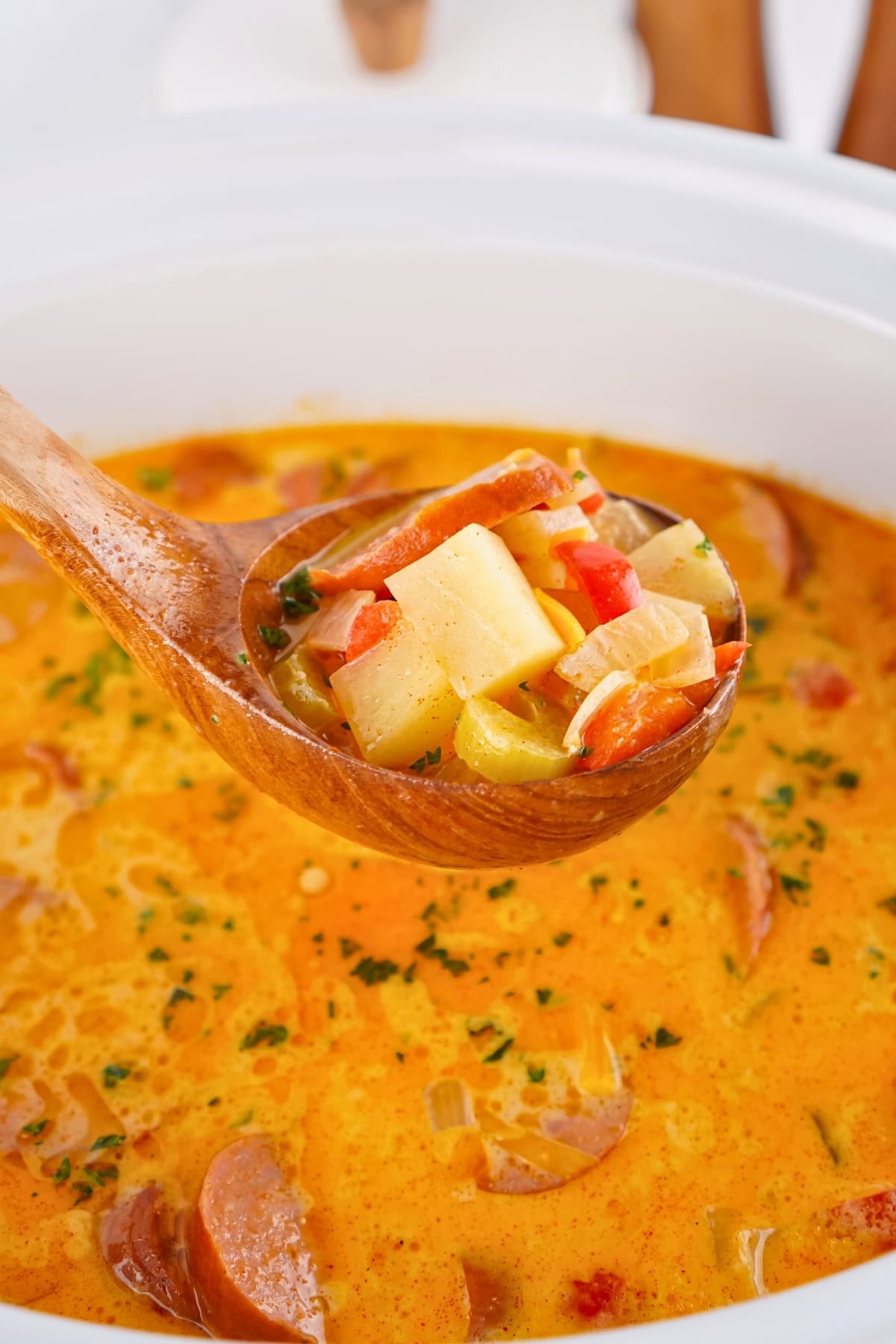 Ladle of creamy sausage and potato soup over a pot, with herbs and vegetables.
