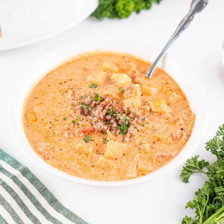 Creamy soup with ground meat, pasta, and herbs in a white bowl with a spoon.