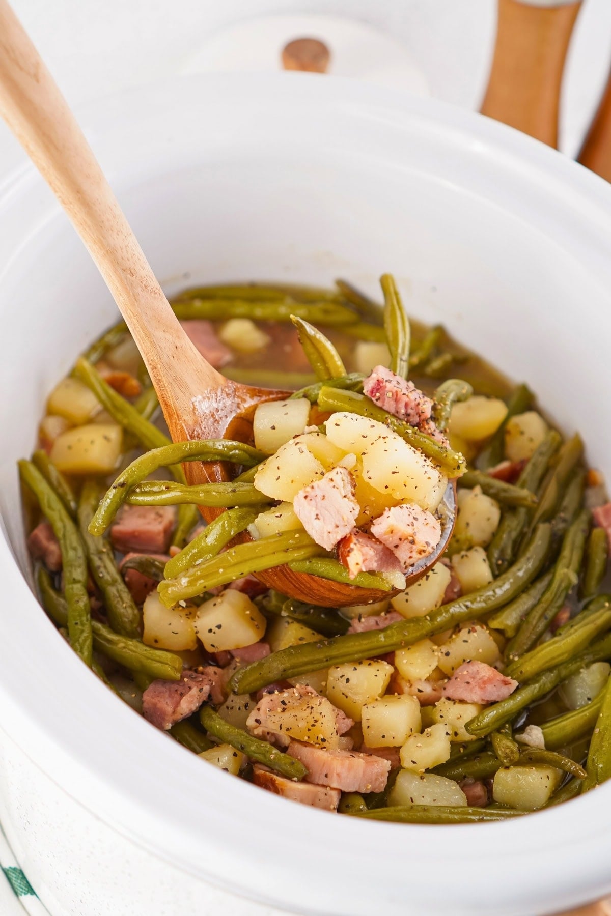 A wooden spoon lifts green beans, potatoes, and ham from a white slow cooker.