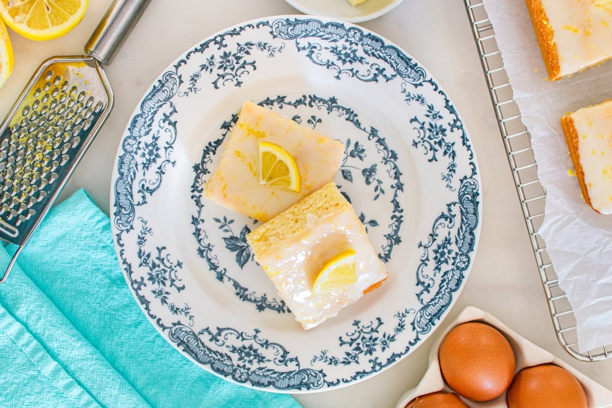 Two lemon bars with glaze and lemon slices on a decorative blue and white plate.