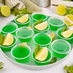 Green Jello shots in cups, topped with lime wedges and mint leaves on a tray.