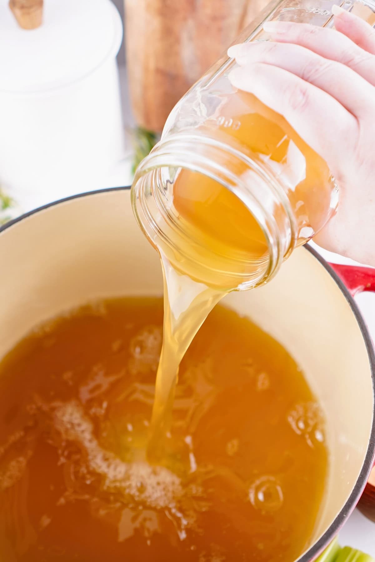 Hand pouring golden broth from a jar into a pot.