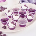 Ten purple gelatin cups, four topped with cream and grape-shaped gummies, on a tray.
