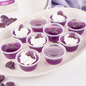 Ten purple gelatin cups, four topped with cream and grape-shaped gummies, on a tray.