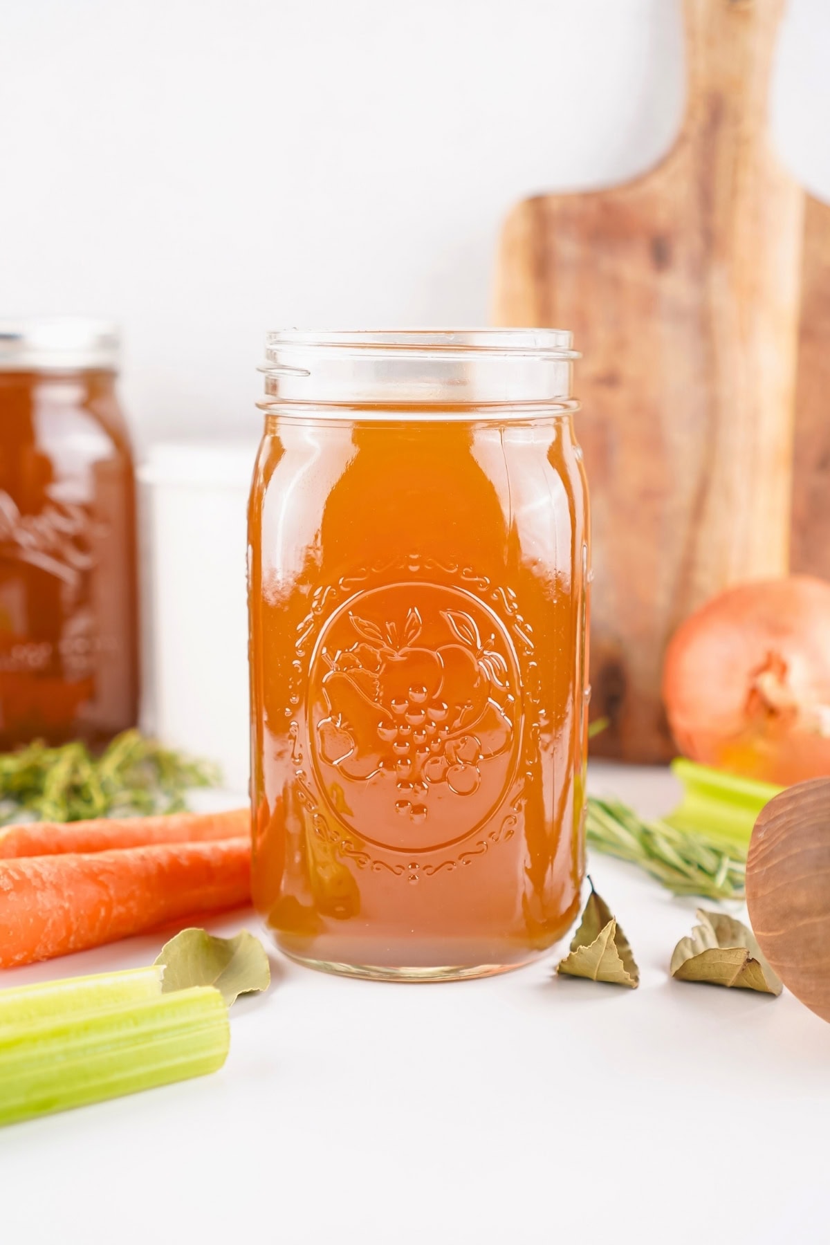 A jar of vegetable broth surrounded by carrots, celery, herbs, and an onion.