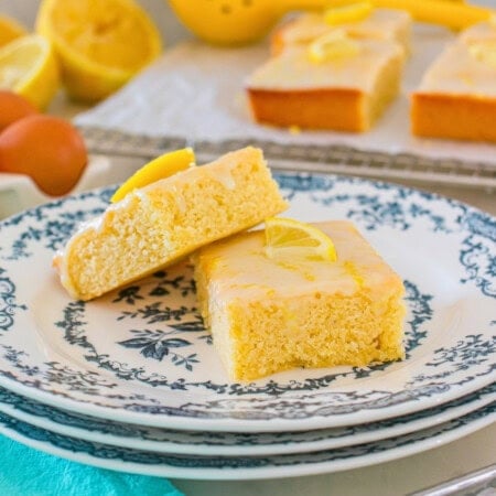 Two lemon cake bars on a plate, topped with icing and a lemon slice.