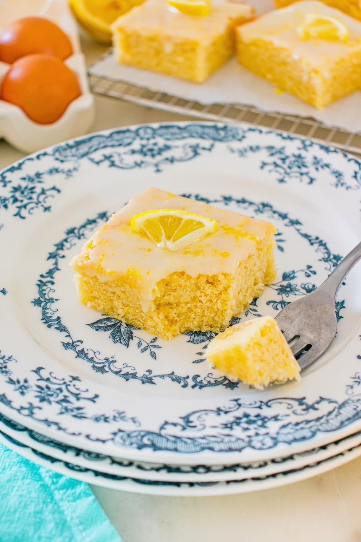 A lemon cake square with icing and a lemon slice on a decorative plate.
