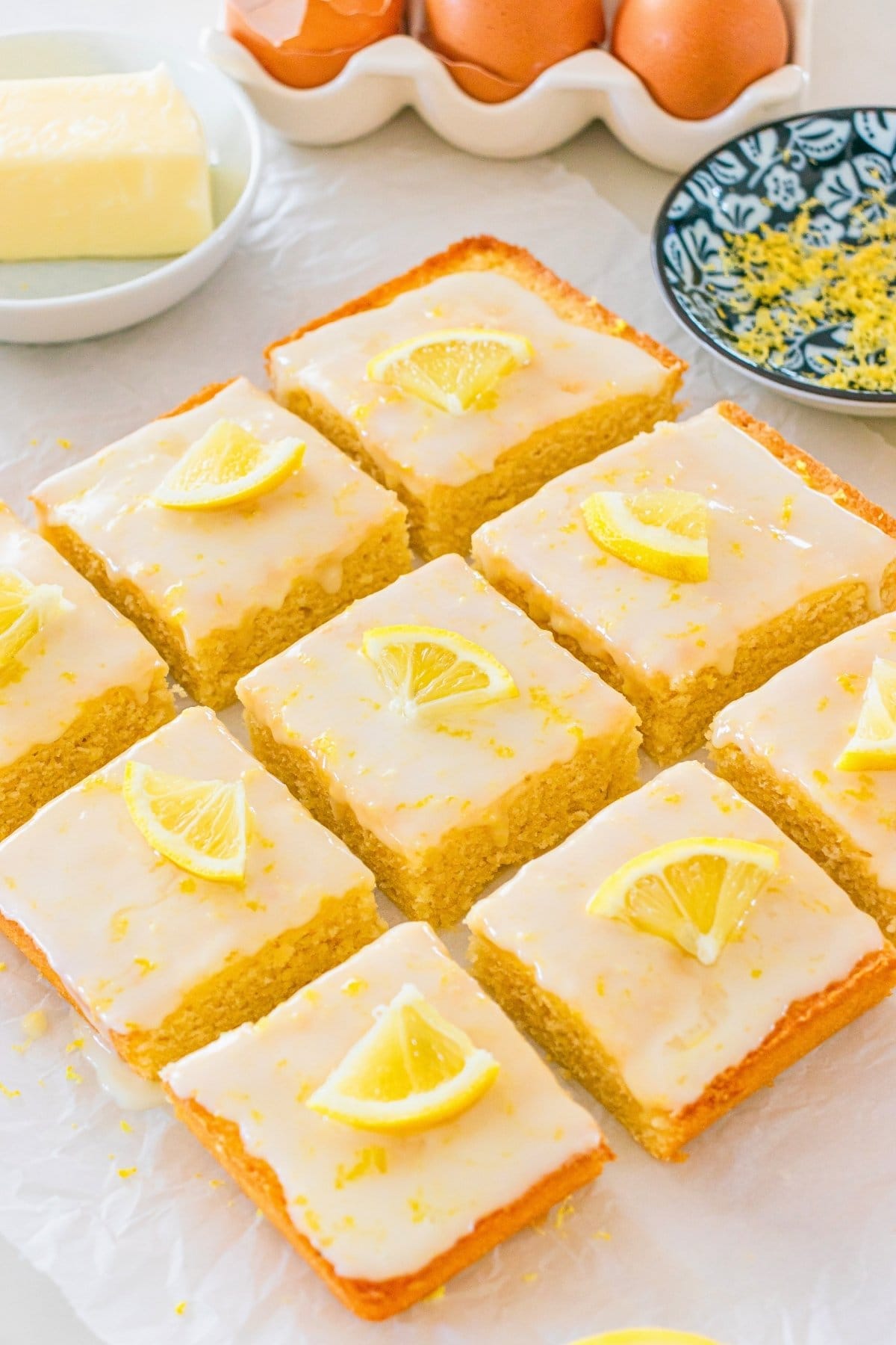 Nine glazed lemon bars topped with lemon slices, with eggs and butter nearby.
