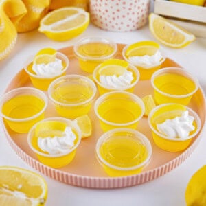 Yellow jello shots with lemon and whipped cream on a pink tray, surrounded by lemons.