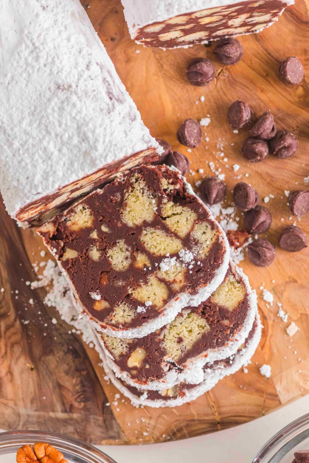 Sliced chocolate salami with powdered sugar on a wooden board, chocolate chips nearby.