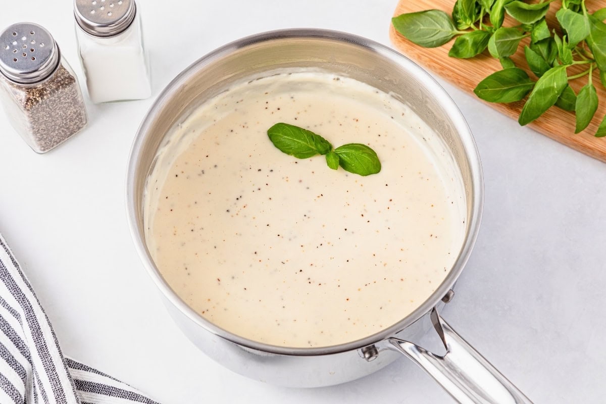Creamy white sauce in a saucepan, garnished with basil leaves, on a white surface.