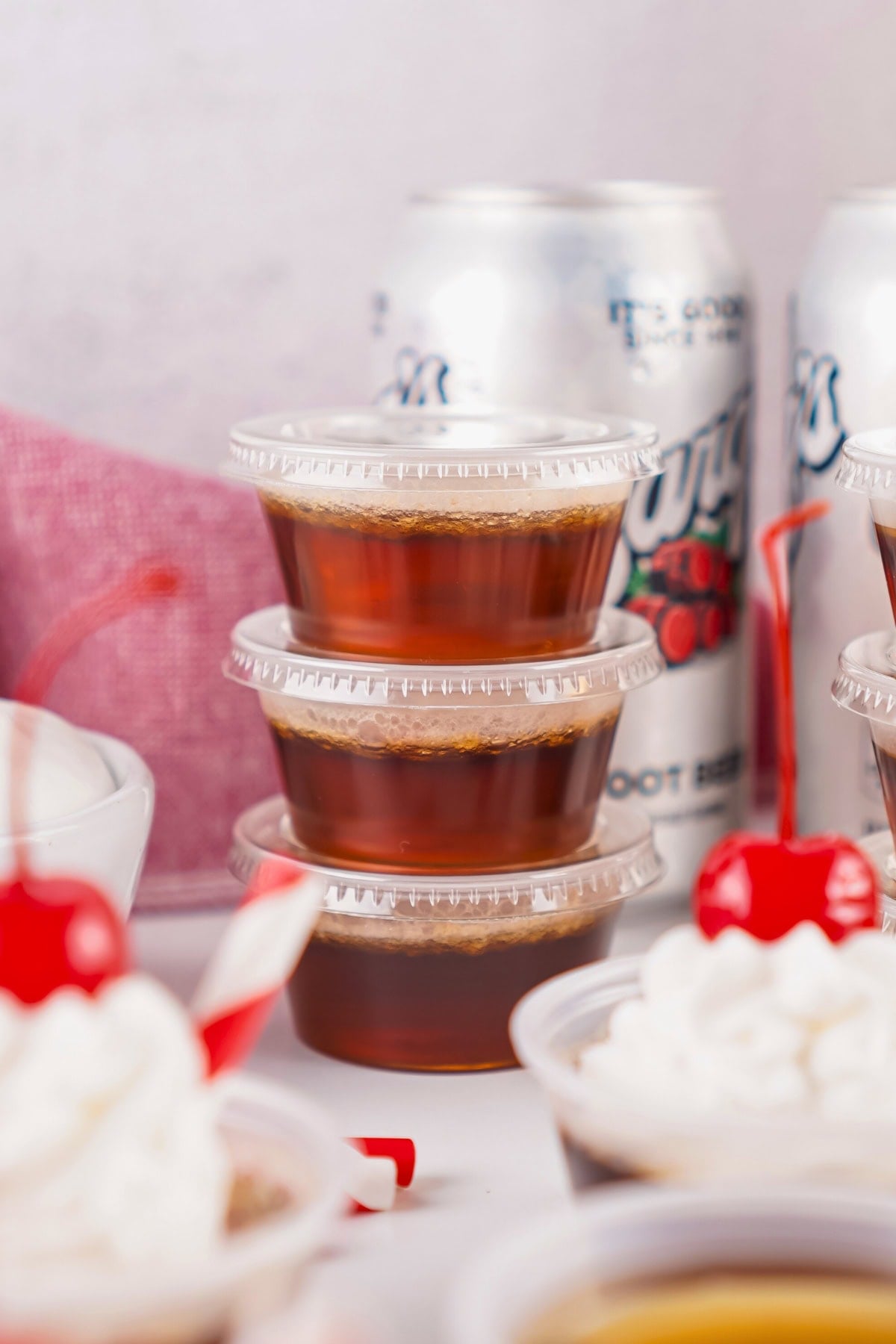 Three stacked plastic cups with dark liquid, with cherries and cans in the background.