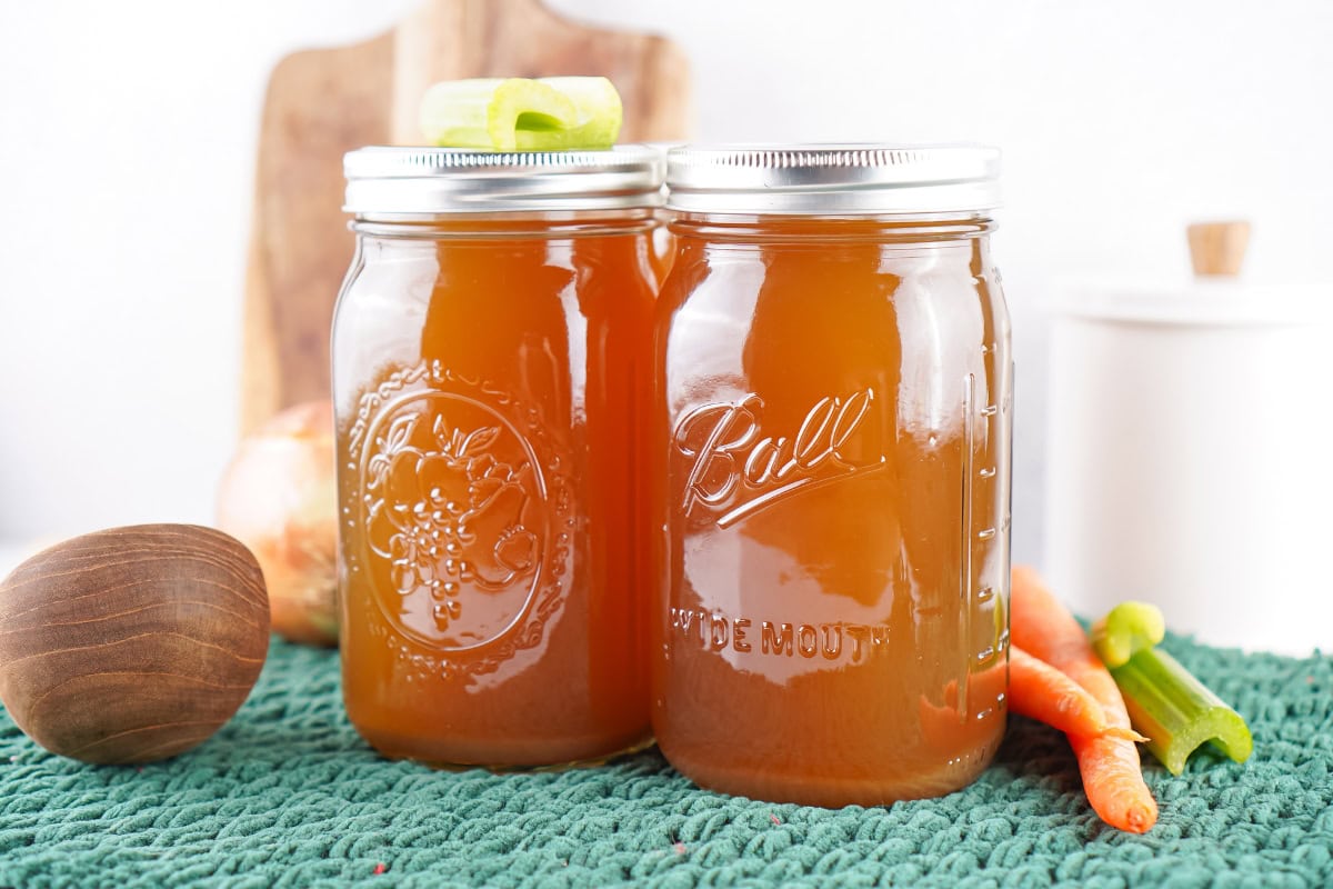 Two glass jars of homemade broth on a green mat with vegetables nearby.