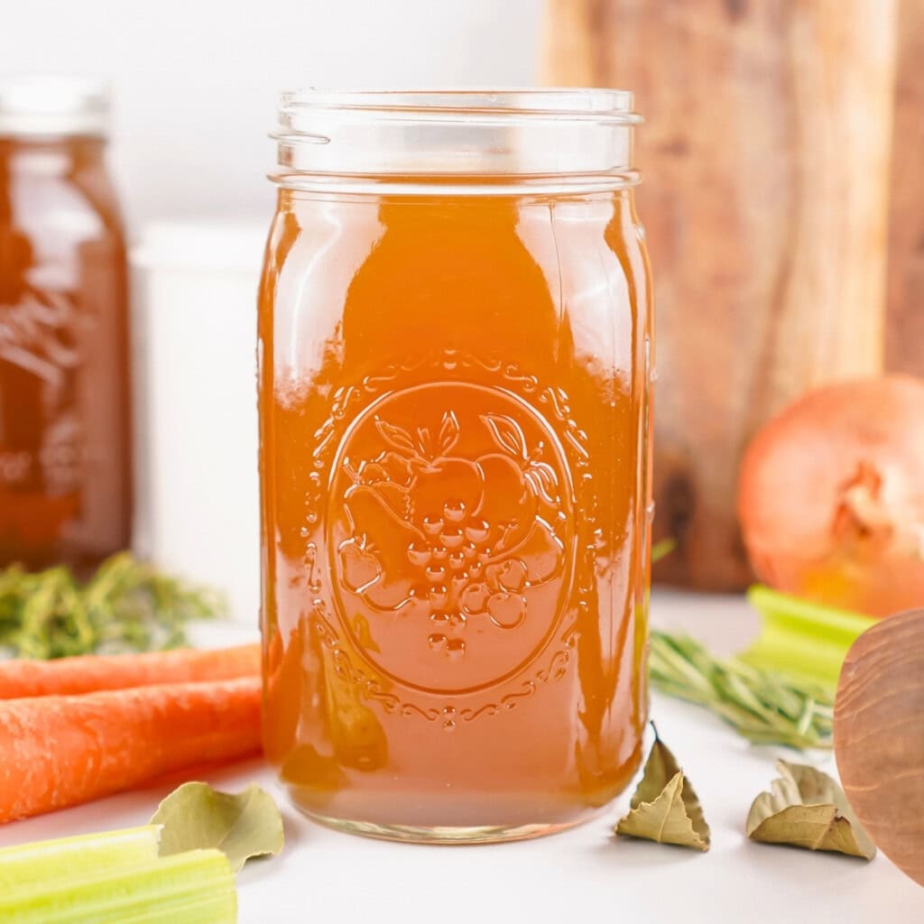 A mason jar of brown broth surrounded by carrots, celery, onion, and herbs.