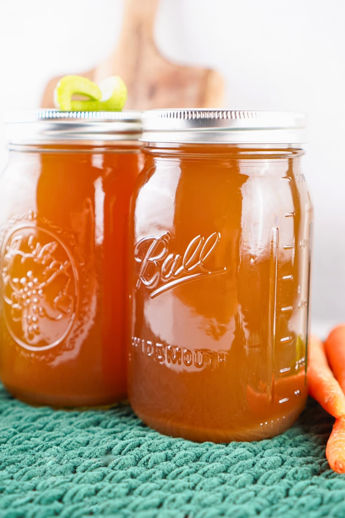 Two jars of homemade broth on a green textured cloth with carrots beside them.