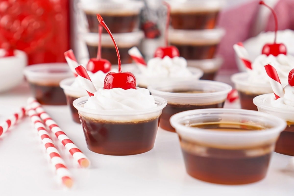Cherry-topped jello shots with whipped cream and striped straws on a white table.