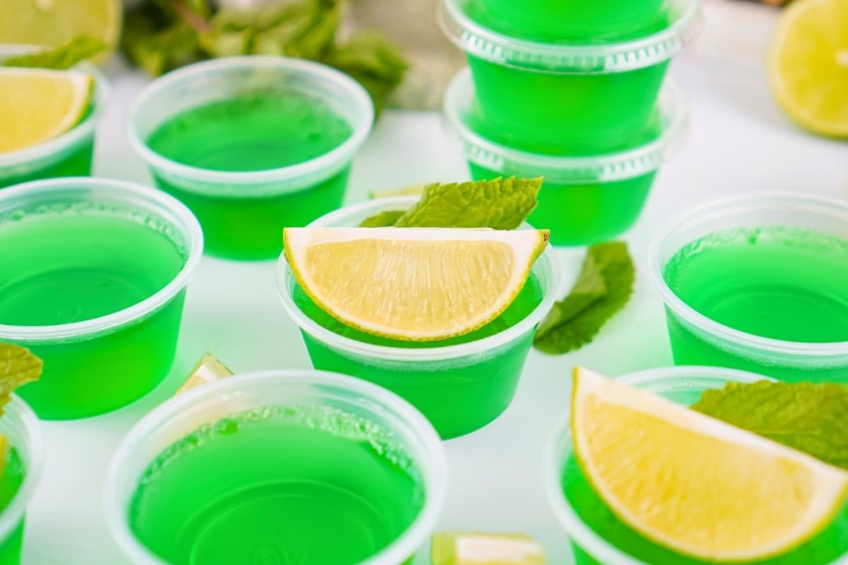 Green gelatin shots in plastic cups, each topped with a lemon wedge and mint leaves.