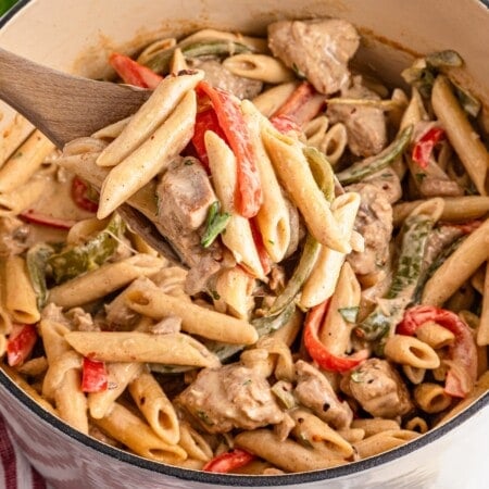 Creamy penne pasta with chicken and bell peppers in a large pot, on a wooden spoon.