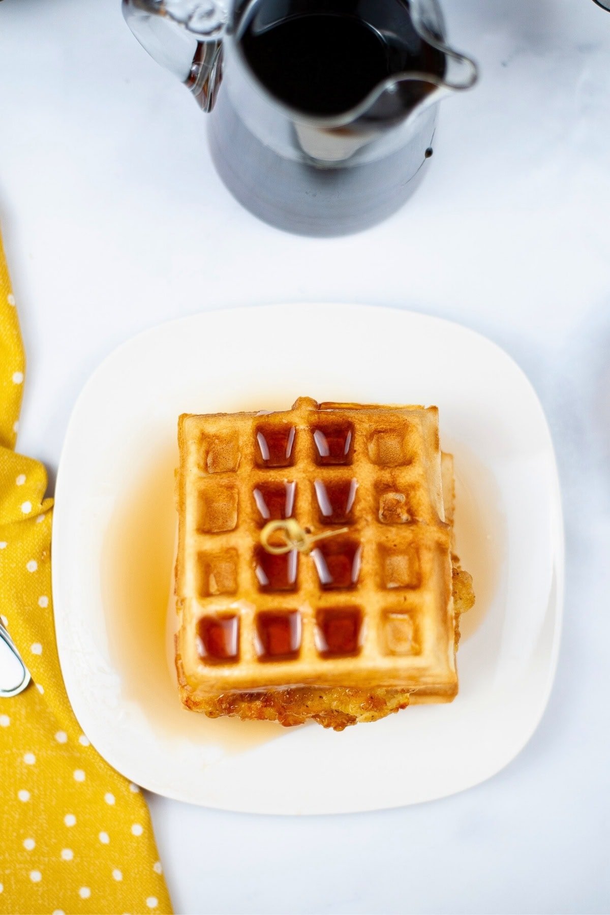 Square waffles with syrup on a white plate, next to a yellow cloth and syrup jug.
