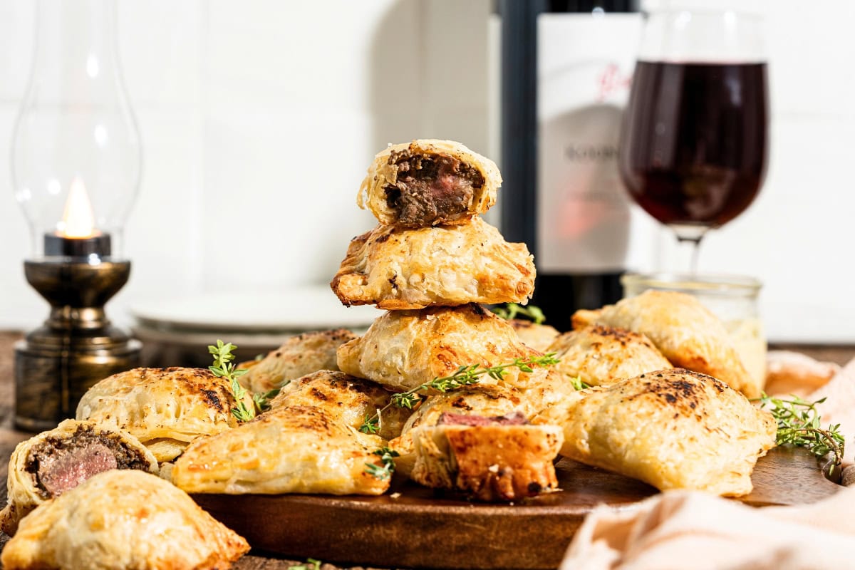 A pile of baked meat pastries on a wooden board with wine and a candle.