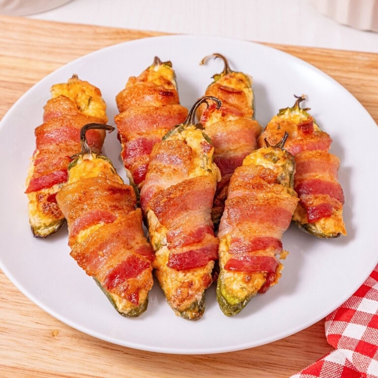 Six bacon-wrapped stuffed jalapeños on a white plate on a wooden surface.