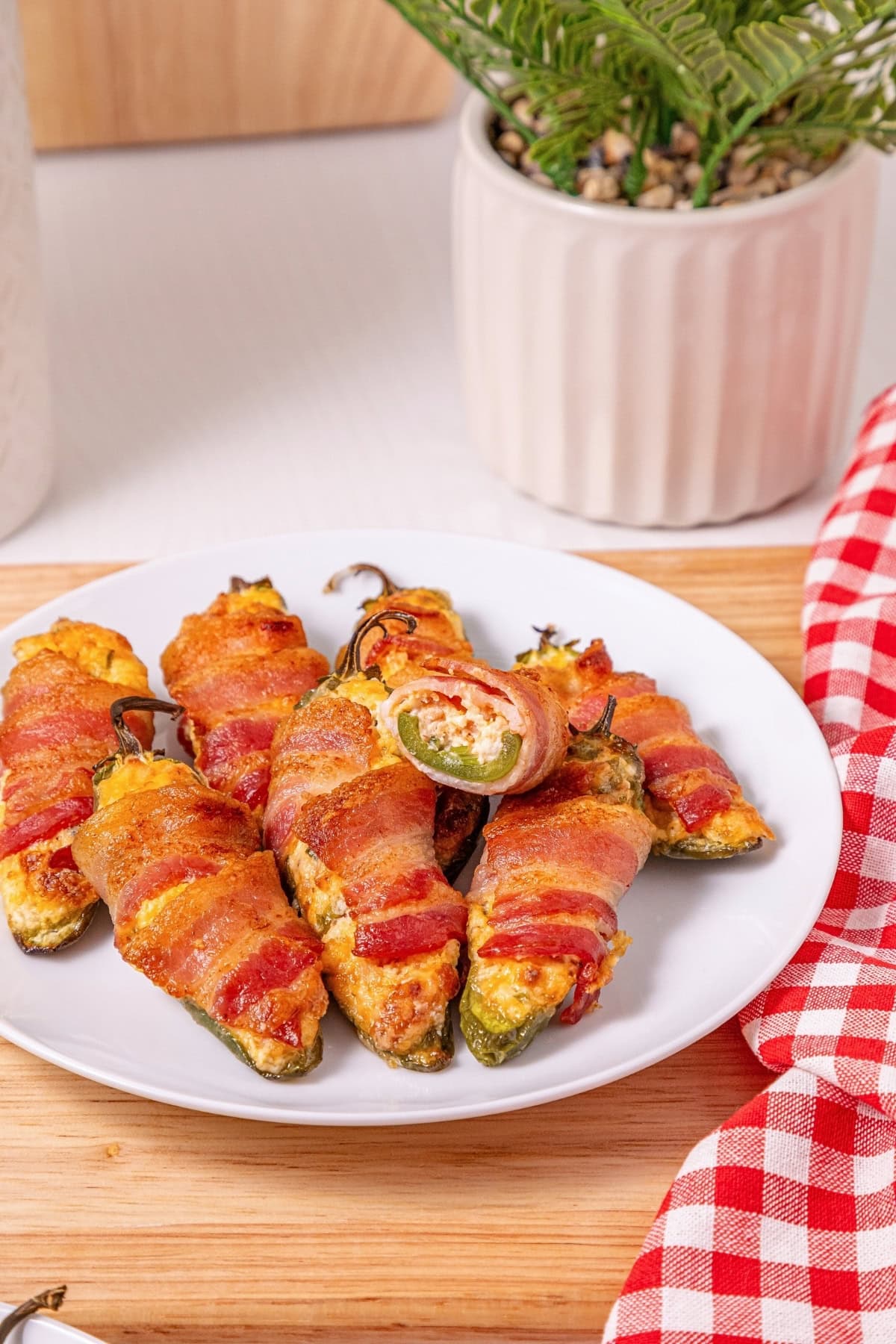 Bacon-wrapped stuffed jalape&ntilde;os on a white plate with a plant and napkin nearby.
